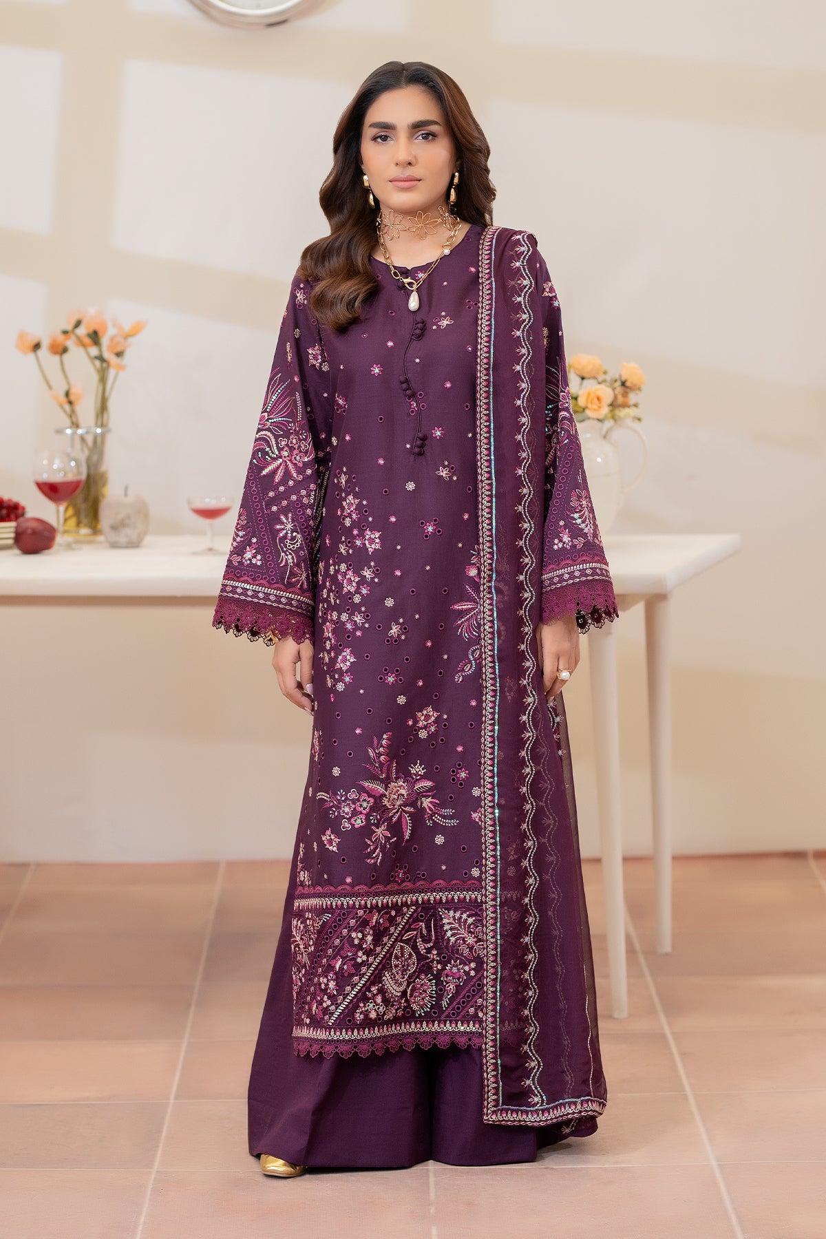 Woman wearing a purple embroidered traditional outfit in a room with a table and flowers.