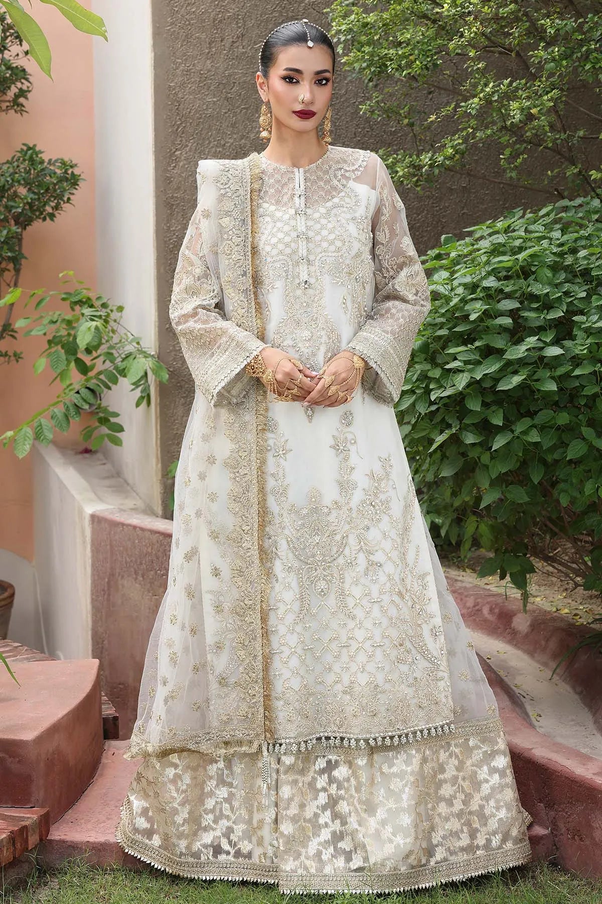 Woman in a white embroidered traditional outfit standing outdoors with greenery.