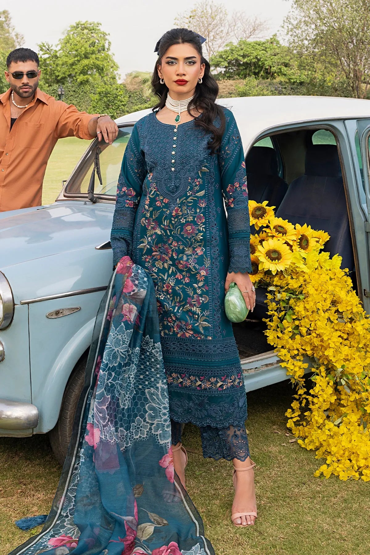 A woman standing beside a vintage car, wearing a teal blue traditional outfit with floral embroidery, including a shirt, dupatta, and trousers.